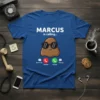 Blue t-shirt with white text "MARCUS is calling..." above a cartoon brown poop wearing sunglasses and phone interface buttons