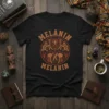 Black t-shirt with "MELANIN MELANIN" text in a circular, tribal-inspired graphic with warm brown and orange tones