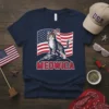 Meowica t-shirt design with a tabby cat wearing a feather headband, standing on rocks with the American flag behind it