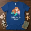 Mermaid Magic text on a blue t-shirt with a cute cartoon mermaid holding a seashell