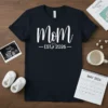 Mom EST 2026 t-shirt with cute baby feet graphic and heart detail, announcing upcoming motherhood in 2026