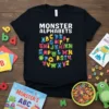 Black t-shirt featuring "MONSTER ALPHABETS" in white text above a circular arrangement of colorful alphabet letters, each accompanied by a