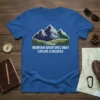 Blue t-shirt with "Mountain Adventures Await Explore & Discover" text and a graphic of majestic mountains and pine trees