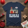My Dad Is A Firefighter text with a cartoon fire station and fire truck design, navy blue t-shirt