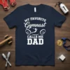 Navy blue t-shirt with white text "My Favorite Gymnast Calls Me Dad" and a silhouette of a gymnast mid-air