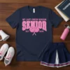 Navy blue t-shirt with "My Last Cheer Season Senior Est. 2026" in pink varsity font, featuring pink pom-poms