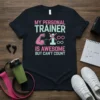 Black t-shirt with text "My Personal Trainer Is Awesome But Can't Count", featuring a pink bicep graphic and mathematical symbols