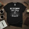 Black t-shirt with white text My Testimony His Glory and an open bible graphic with a cross and sunburst