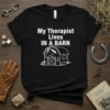 Black t-shirt with white text "My Therapist Lives IN A BARN" and a graphic of a horse in a barn