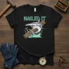 Nailed It text with a cartoon shark hammering a nail into wood, on a black t-shirt
