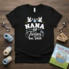 Black t-shirt with "Nana Of Twins Est. 2025" in white and blue text, featuring stork graphics