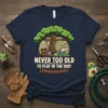 T-shirt with text "Never Too Old To Play In The Dirt" featuring a tree, shovel, lawn mower, and boots