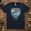 Navy blue t-shirt with "Ocean Catch Est 1980" text and a jumping fish graphic in a shield, surrounded by waves