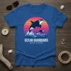 Orca whales jumping waves with sunset graphic, text reads OCEAN GUARDIANS WAVE RIDERS, on a blue t-shirt