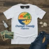 White t-shirt featuring a circular design with palm trees, a sunset over the ocean, and the text "Orange County California"