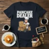 Pancake Dealer T-Shirt featuring a cartoon chef with a cart full of pancakes and money bags, with bold white text