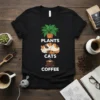 Black t-shirt with "PLANTS CATS COFFEE" text and graphics of a potted plant, a sleeping cat, and a coffee cup