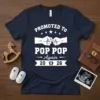 Promoted to Pop Pop Again 2026 text and graphic design on a navy t-shirt, featuring fists bumping and baby footprints
