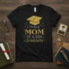 Proud MOM OF A 2026 Graduate text with a gold graduation cap graphic on a black t-shirt