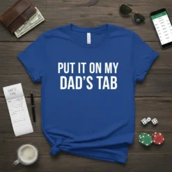 Blue t-shirt with white bold text reading "PUT IT ON MY DAD'S TAB". The shirt is laid flat on a