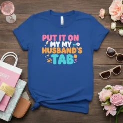 Blue t-shirt with bold text "PUT IT ON MY MY HUSBAND'S TAB" in pink, orange, and teal, featuring shopping-related icons