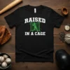 Baseball player swinging bat in a cage graphic with "RAISED IN A CAGE" text on a black t-shirt