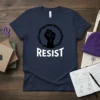 Navy blue t-shirt with bold white text "RESIST" and a black graphic of a raised fist inside a thorny circle
