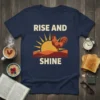 RISE AND SHINE t-shirt with a graphic of a rooster standing before a rising sun and hills