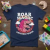 Roar Magiic Unicorn Power text with a pink T-Rex wearing a unicorn horn, surrounded by a rainbow and clouds on