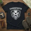 Black t-shirt featuring a white graphic of a Sabertooth skull within a shield, with mountains and tribal patterns. Includes the