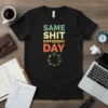 Black t-shirt with "SAME SHIT DIFFERENT DAY" in retro font and circular icons representing daily tasks