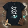Black t-shirt with white text "Sand. Surf. Shark Teeth." and a wave graphic, featuring shark teeth scattered around