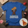 Sasquatch character holding a slice of pizza above its head with "Sasquatch Loves Pizza" text below on a blue t-shirt
