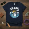 Shark Life t-shirt design with a cool shark wearing sunglasses surfing on waves under a sun graphic