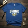 Blue t-shirt with the word SHINE in bold white letters and a golden sun graphic below, surrounded by stars