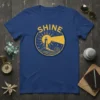 Yellow lighthouse graphic with "Shine" text on a navy blue t-shirt, symbolizing guidance and hope