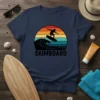 Skimboarder silhouette riding a wave against a retro sunset on a navy blue t-shirt