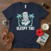 Sleepy Tiki text design with a cartoon tiki character relaxing in a hammock between palm trees, navy blue t-shirt