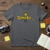 Grey t-shirt with yellow "Smile_" text, a smiling sun graphic, and a camera lens icon