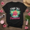 Black t-shirt with "Stand Tall And Be Fabulous" text, featuring a pink flamingo on a surfboard with tropical leaves and
