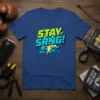 Stay Srng! quote in bold green and blue typography on a blue t-shirt, with brick wall and lightning graphic