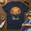 Retro sunset graphic with surfboards and text "SURF AT Beach" on a navy blue t-shirt