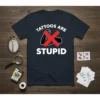 Dark navy t-shirt with white text "TATTOOS ARE STUPID" and a red cross over a muscular arm graphic