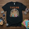 Black t-shirt with "Taylor" in orange text above a colorful castle with cats dressed as royalty, surrounded by flowers