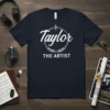 Navy t-shirt with white text "Taylor THE ARTIST" in a circular design with laurel leaves and sound wave graphic