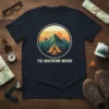 The Adventure Begins quote on a navy t-shirt with a vintage camping scene graphic featuring mountains, tent, and campfire