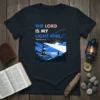 Christian t-shirt with text "The Lord Is My Light And Psalm 27:12" above a lighthouse illustration with a person standing