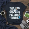 Navy t-shirt with white text: "THIS IS MY SOCCER PAJAMA SHIRT." Features graphics of a soccer ball, goal, person holding