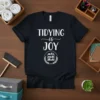 Black t-shirt with white text "TIDYING is JOY" and a graphic of a bookshelf within a laurel wreath