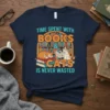 Time Spent With Books & Cats Is Never Wasted quote on a navy t-shirt, featuring cartoon cats and a bookshelf